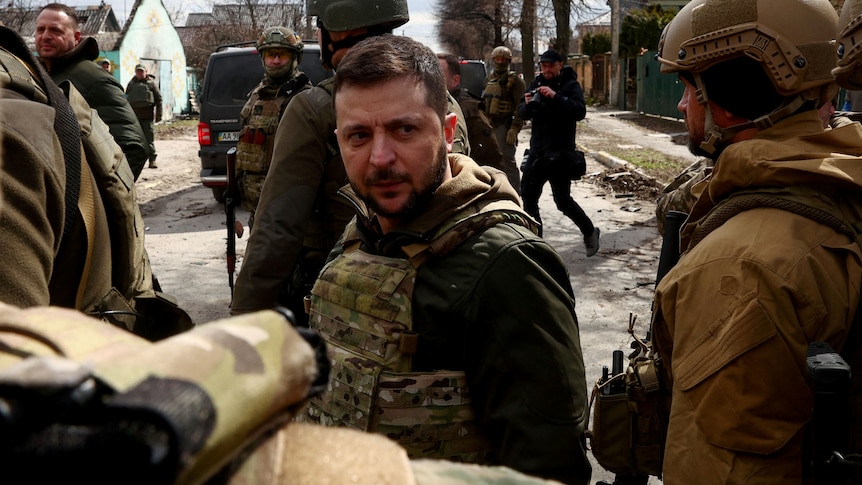 Mr Zelenskyy stands in protective gear but no helmet on the streets of Bucha, with other soldiers and civilians in the frame.