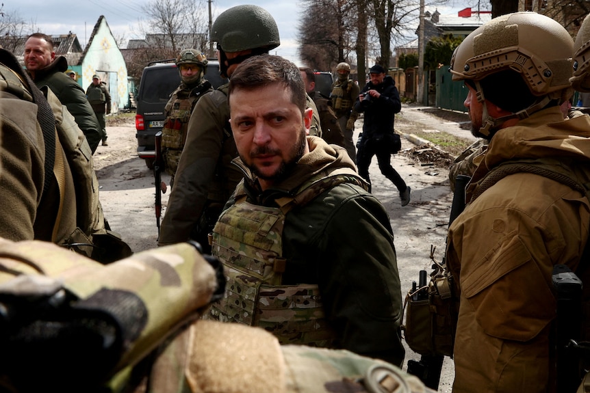 Mr Zelenskyy stands in protective gear but no helmet on the streets of Bucha, with other soldiers and civilians in the frame.