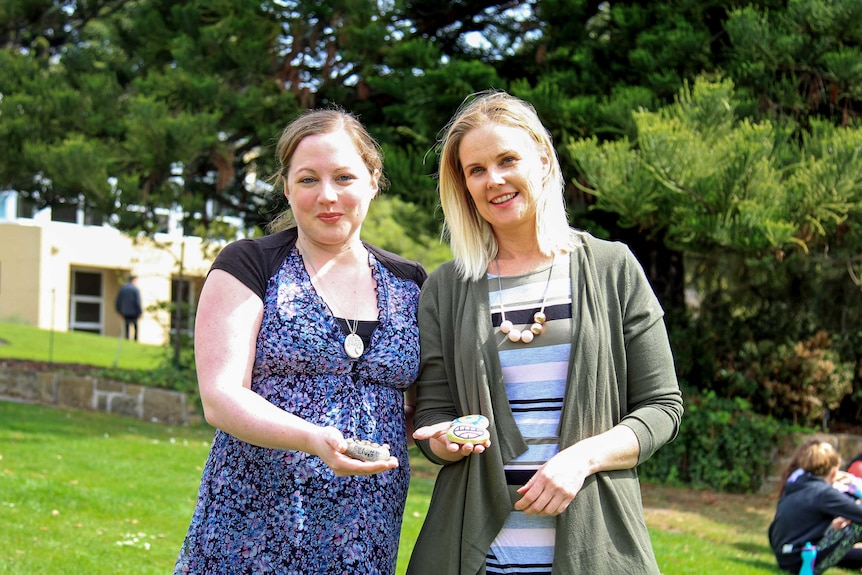 Two women holding painted rocks and smiling in a green garden