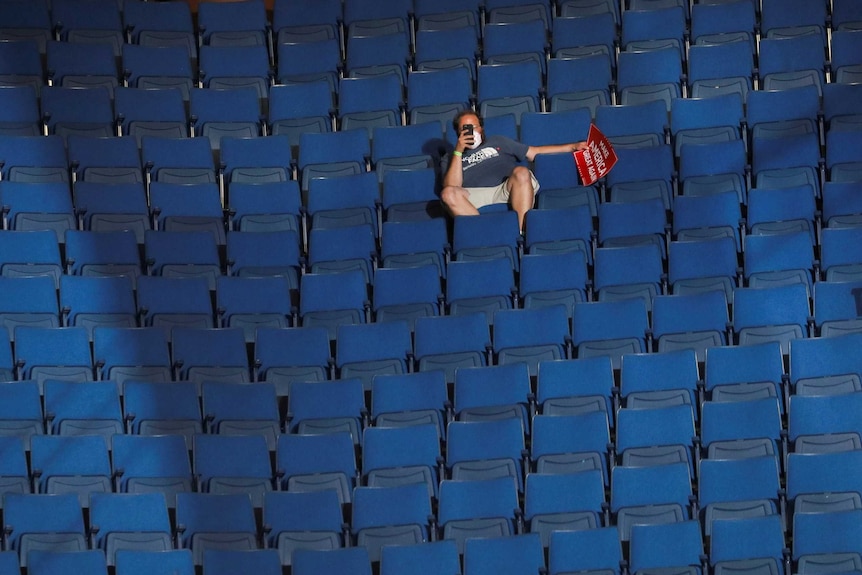 A man sits in an empty row of seats looking at his phone, while holding a 'make America great again' sign