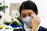 A woman with black hair and wearing a mask holds a cloth to her face as she cries.