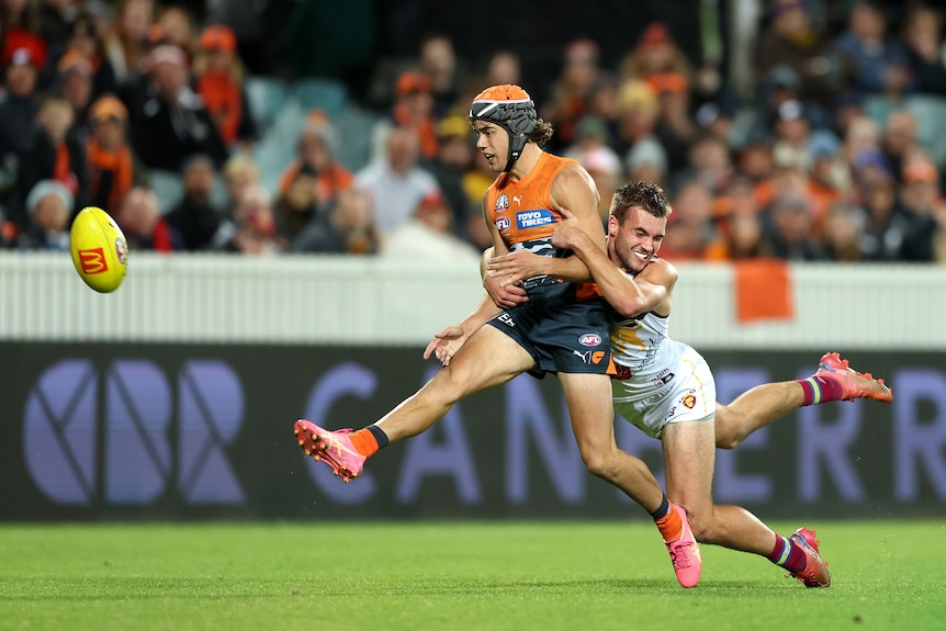 A GWS player wearing orange and black headgear extends his foot after kicking the ball towards goal as he is tackled. 