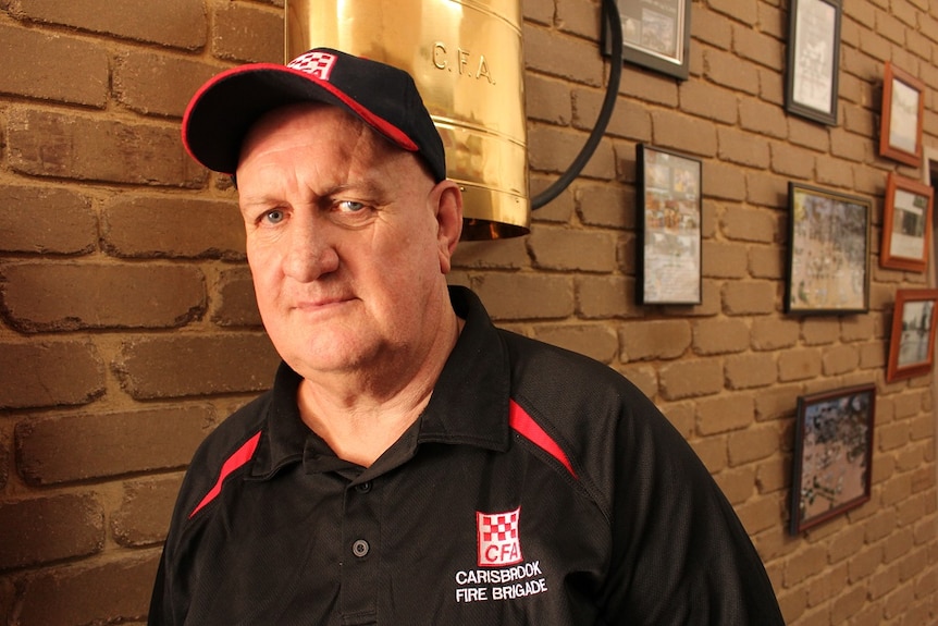 A man with a black and red polo shirt and cap stands in front of a brick wall with framed photographs.