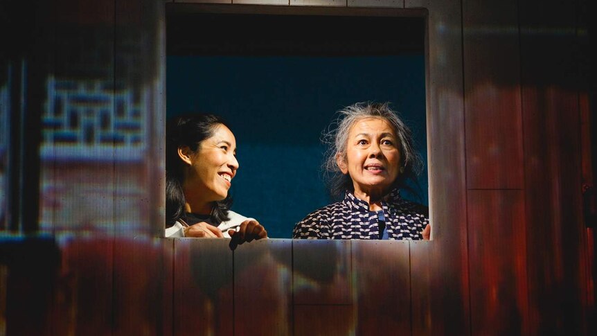 Alice Keohavong (left) and Amanda Ma (right) look out of a window of a building on stage with wooden panels projected on it.