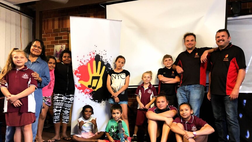 Nine indigenous kids of different ages and their teacher grouped around their choir poster with an Aboriginal flag-based logo.