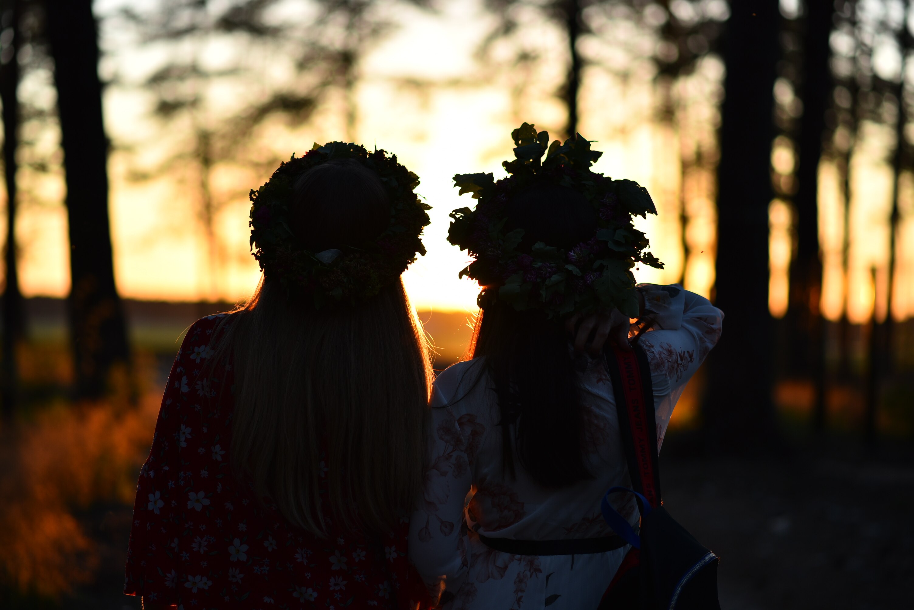 Silhouette of two young people wearing traditional wreaths looking to sunset through forest treesduring the midsummer holiday