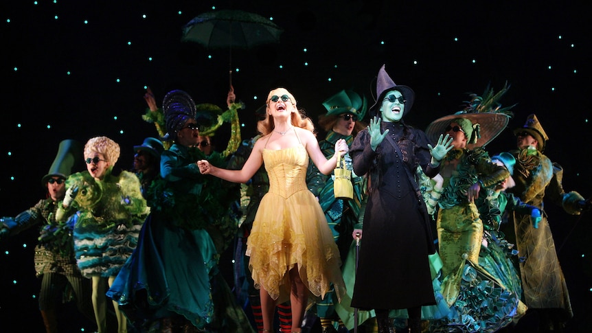 On stage a blonde woman in a champagne dress sings beside brunette woman with green skin in dark cloak and witch's hat.
