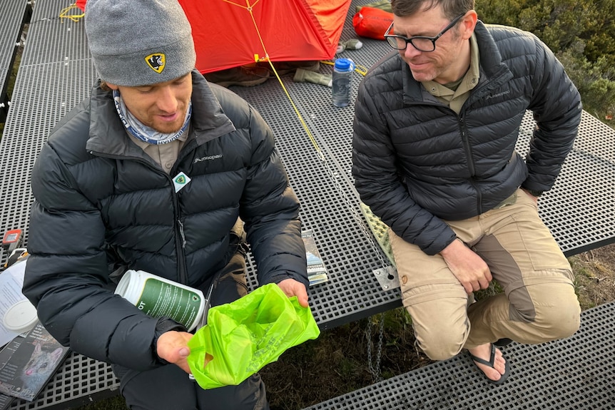 Two men sitting on a national parks camping platform, one holding a poo pot and a corn starch bag.