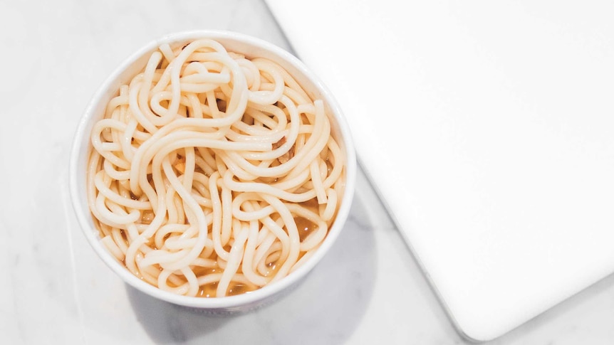 A cup of noodles sits beside a laptop computer, with two wooden chopsticks nearby. A quick meal.