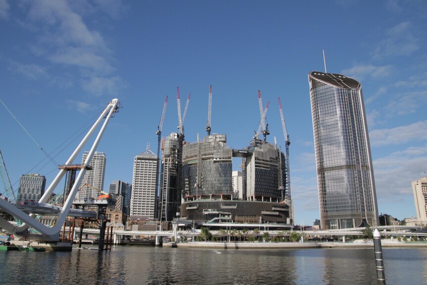 A cityscape with numerous cranes mingled with skyscrapers, as seen from across a river.