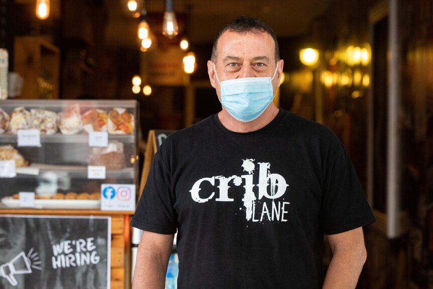 A mid shot of a man wearing a face mask and black t-shirt while posing for a photo in a coffee shop.