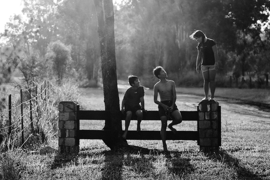 Black and white photo of William and Heetae sitting on a fence, Claire standing.