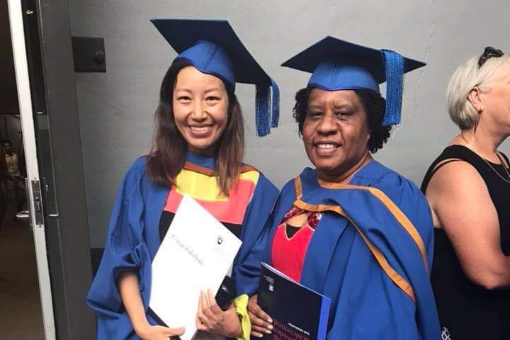 Nancy Wii wearing a blue cap and gown stands holding a certificate next to a woman wearing the same cap and gown.