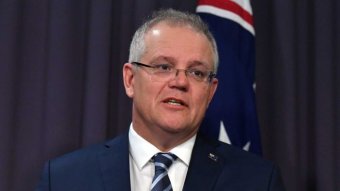 Scott Morrison looks to the right as he stands in front of a blue background mid-speech. An Australian flag is also behind him.