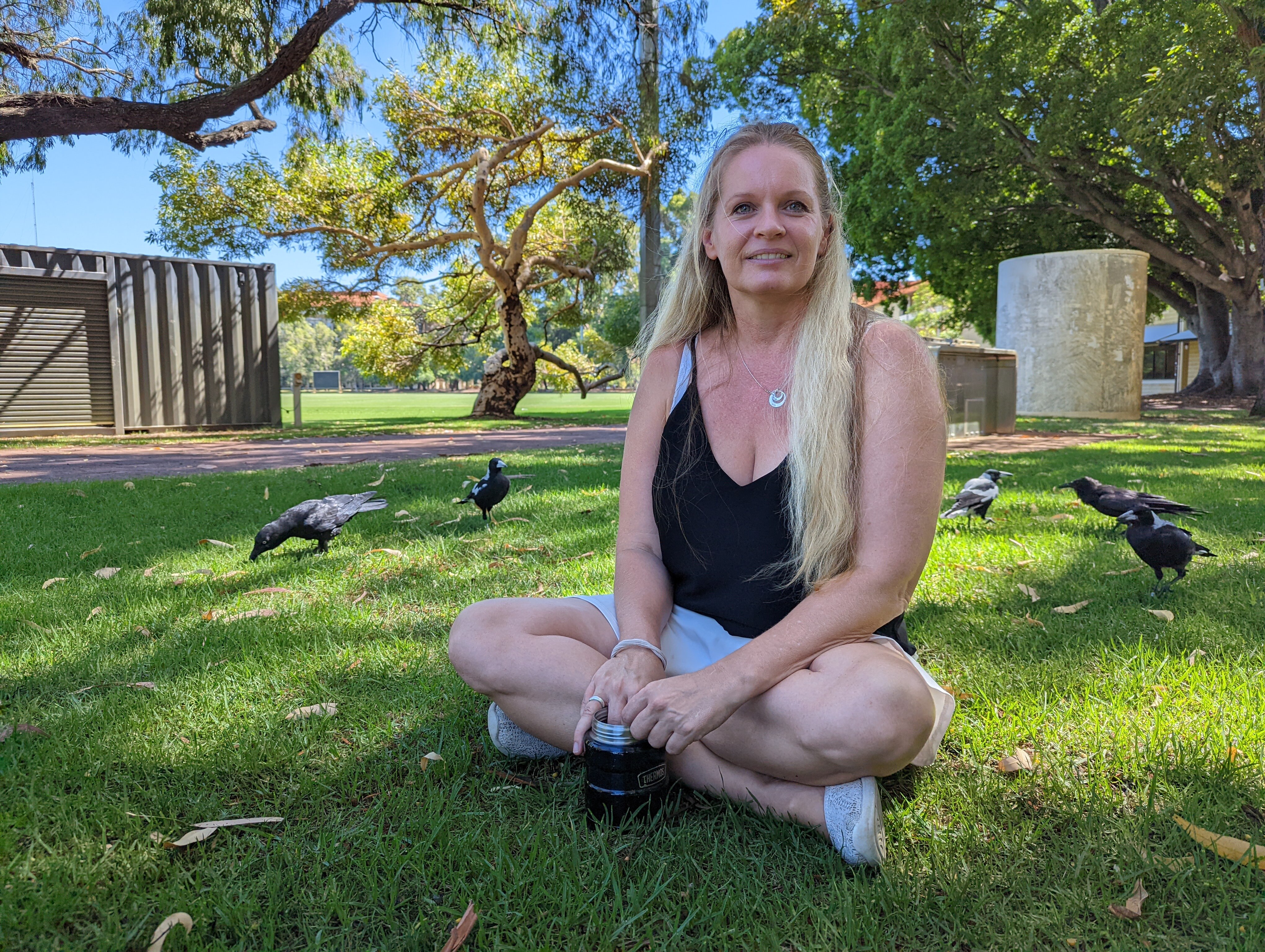 Woman with long blond hair in singlet sits on grass with crows and magpies around her