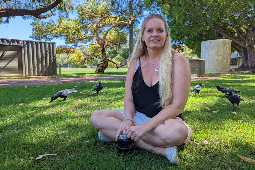 Woman with long blond hair in singlet sits on grass with crows and magpies around her