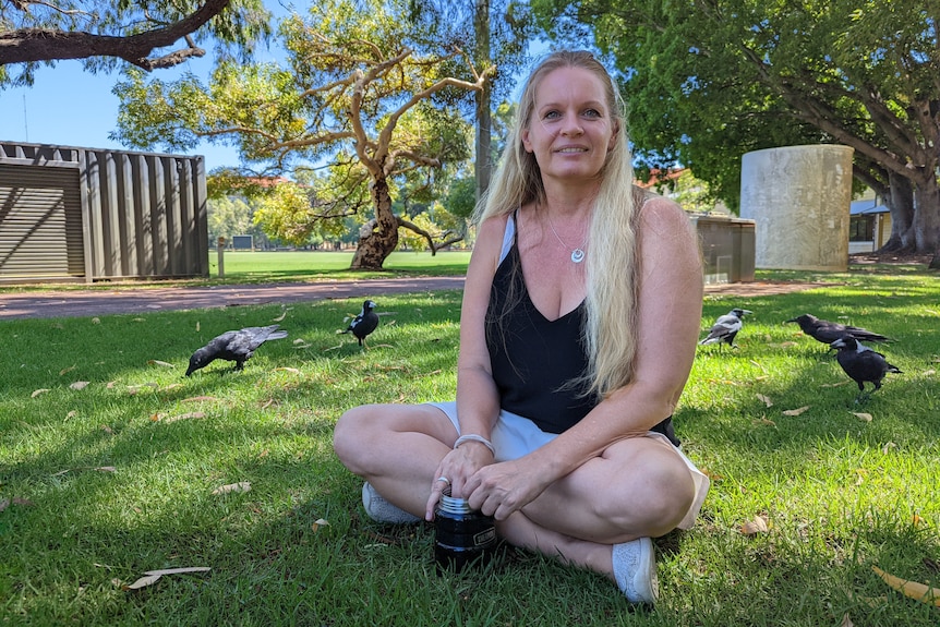 Woman with long blond hair in singlet sits on grass with crows and magpies around her