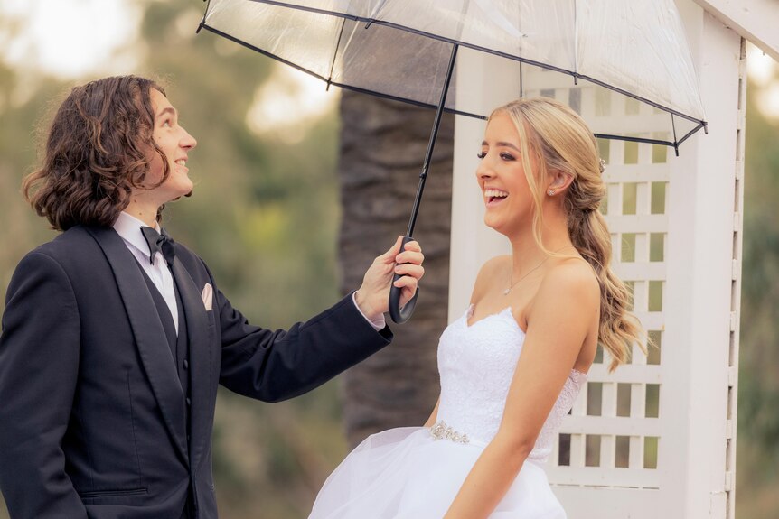 A young man and woman, both in formal attire, smile while standing underneath an umbrella.