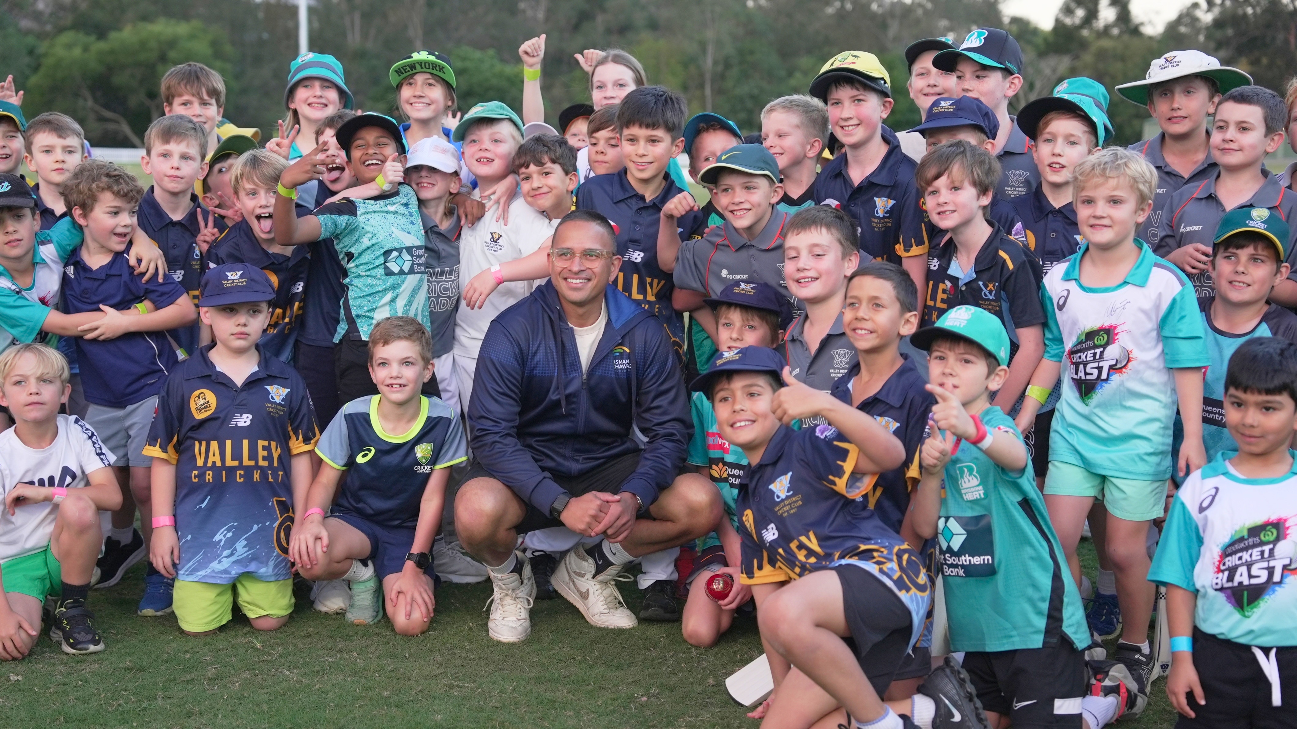 Cricketer Usman Khawaja kneels down in front of a large group of excited children.