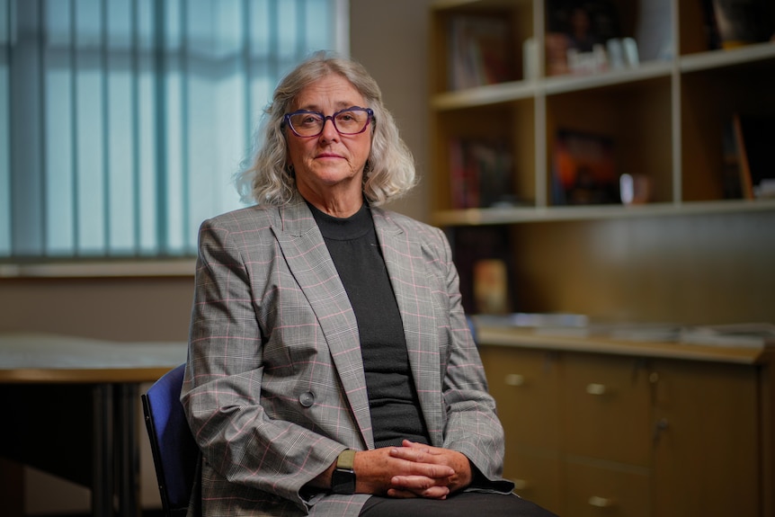 A woman wearing glasses sits in a room in front of a book shelf, looking at the camera 