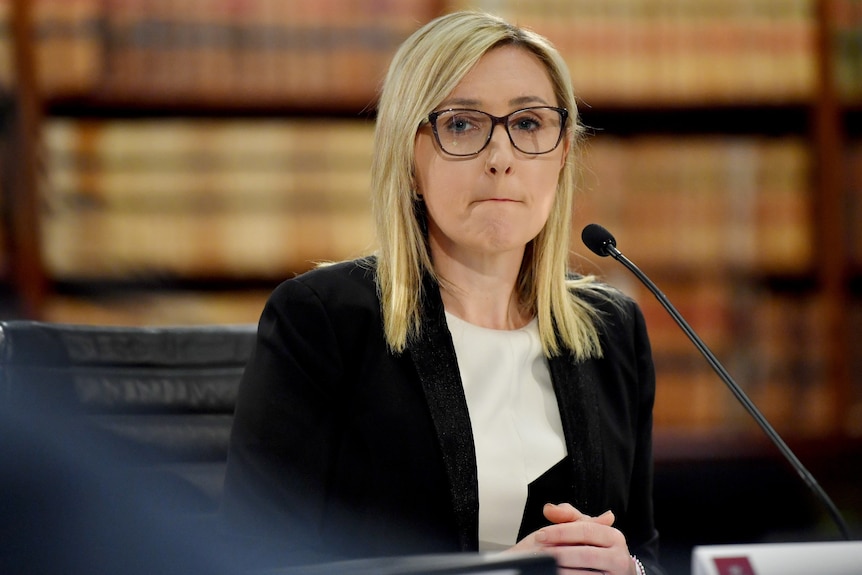 A blonde woman wearing glasses sits behind a desk and microphone.