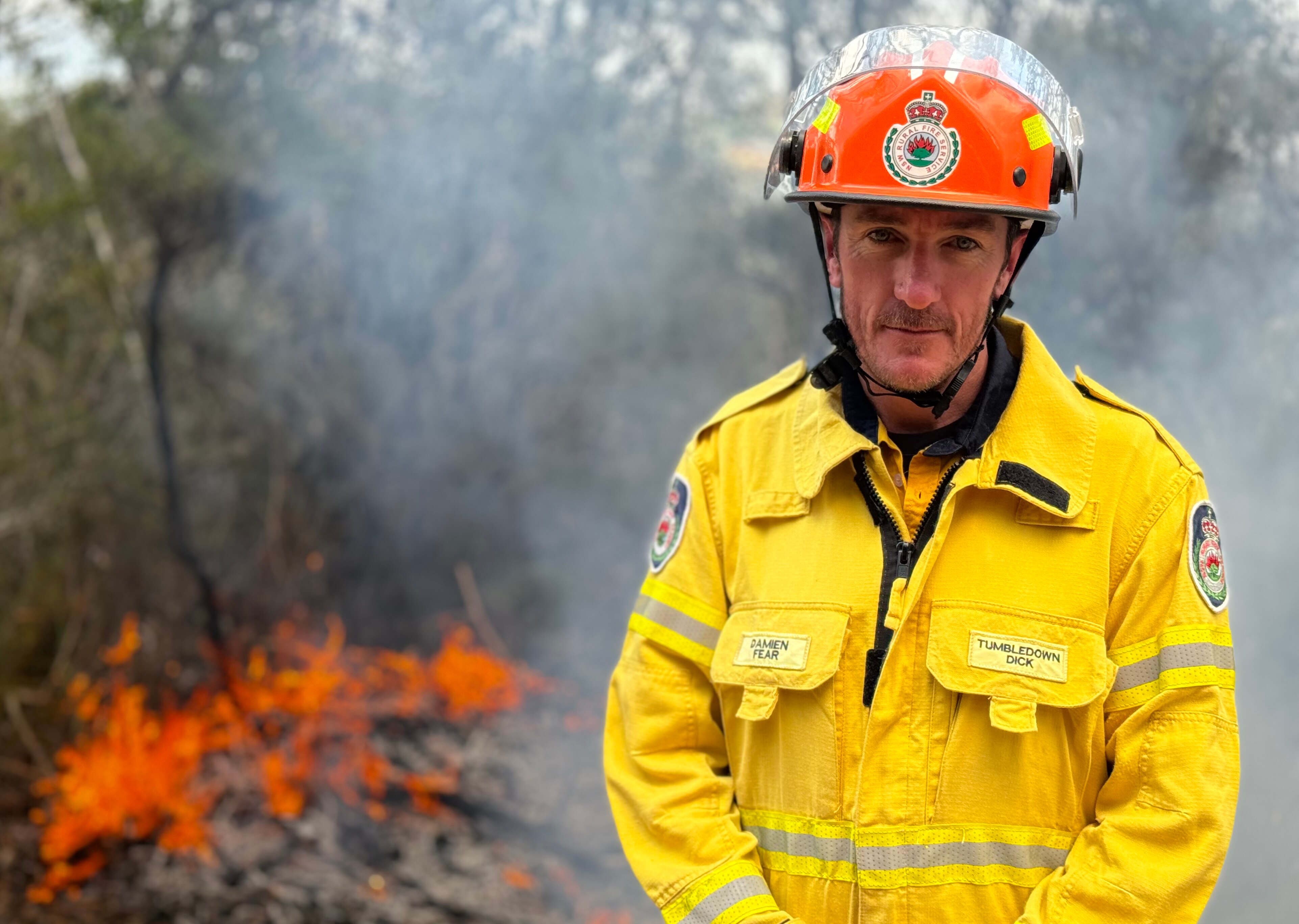 Damien Fear looks intensely at the camera while a fire burns behind him.