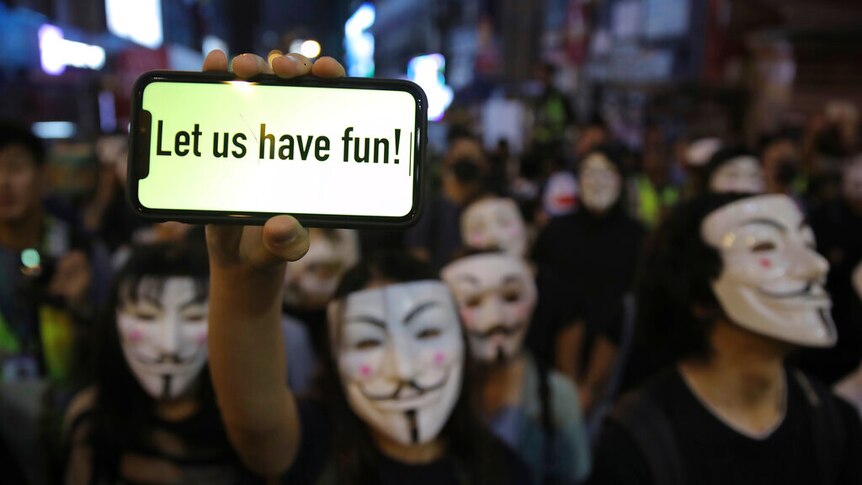 A person holds a smartphone in a crowd people wearing Guy Fawkes masks which reads 'Let us have fun!'.