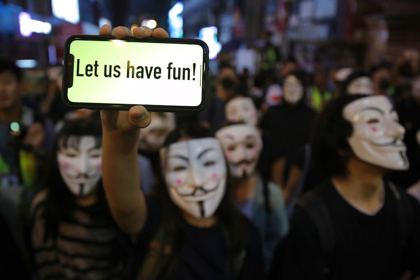 A person holds a smartphone in a crowd people wearing Guy Fawkes masks which reads 'Let us have fun!'.