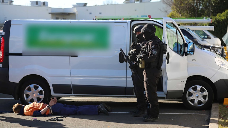 A man lies on the ground in handcuffs stood over by armed police