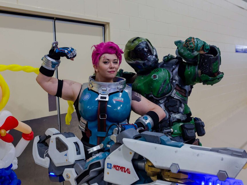 A woman poses flexing her arm while holding a big gun as character from Overwatch. Man in green suit behind her.