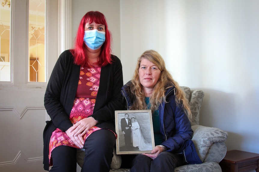 Two middle-aged women sit on a chair, one of them is holding a framed photograph of their mum on their wedding day