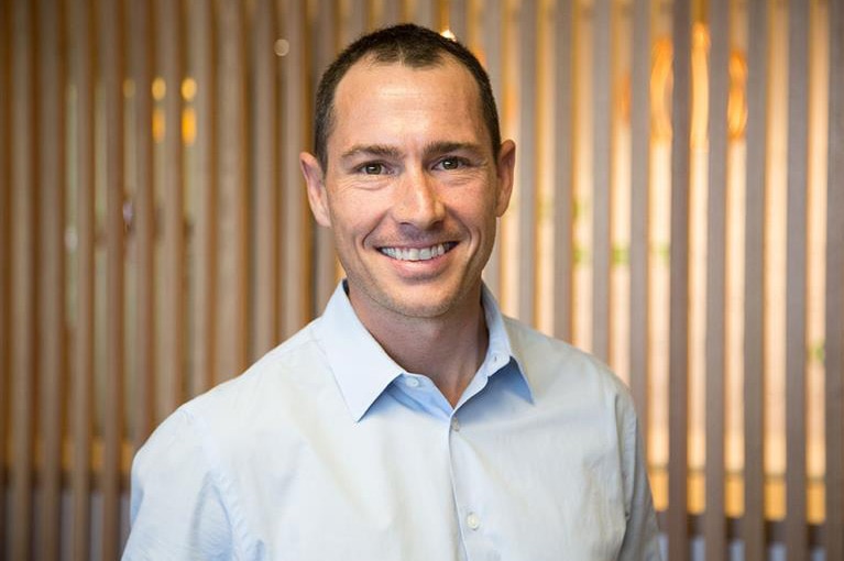 A man wearing a white business shift poses for a photo indoors smiling.