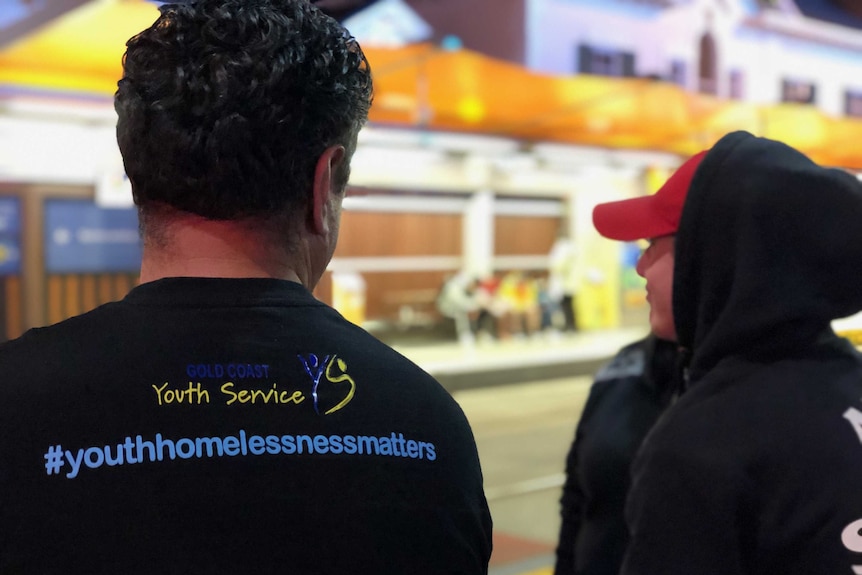 A man wearing a t-shirt that says Gold Coast Youth Service talks to a young man wearing a red cap.