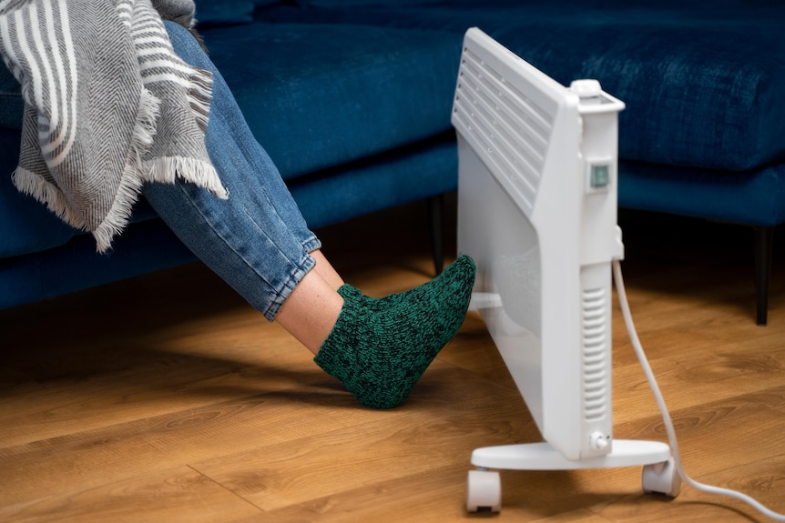 A portable electric heater on the floor with a person's feet warming up next to it