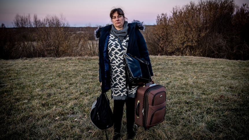 A woman in a big parka holding two bags stands in a field at sunset 