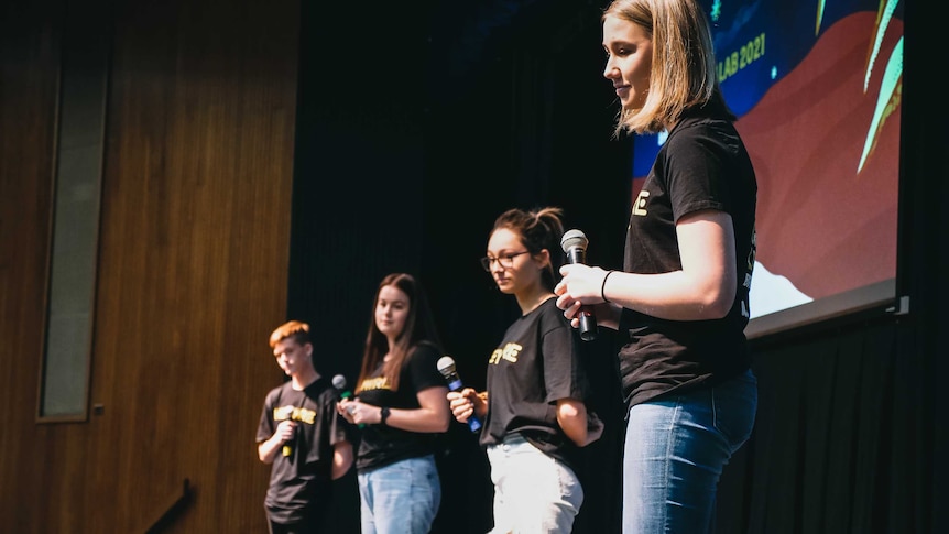 Four young people on stage holding microphones, they are all wearing black t-shirts with the word 'Heywire' written on them