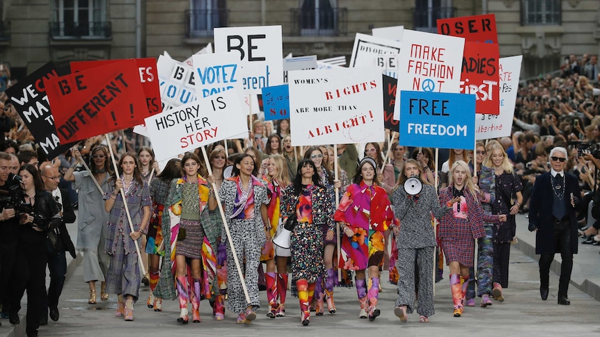 A large group of models walk down a runway holding placards with various slogans and words on them.