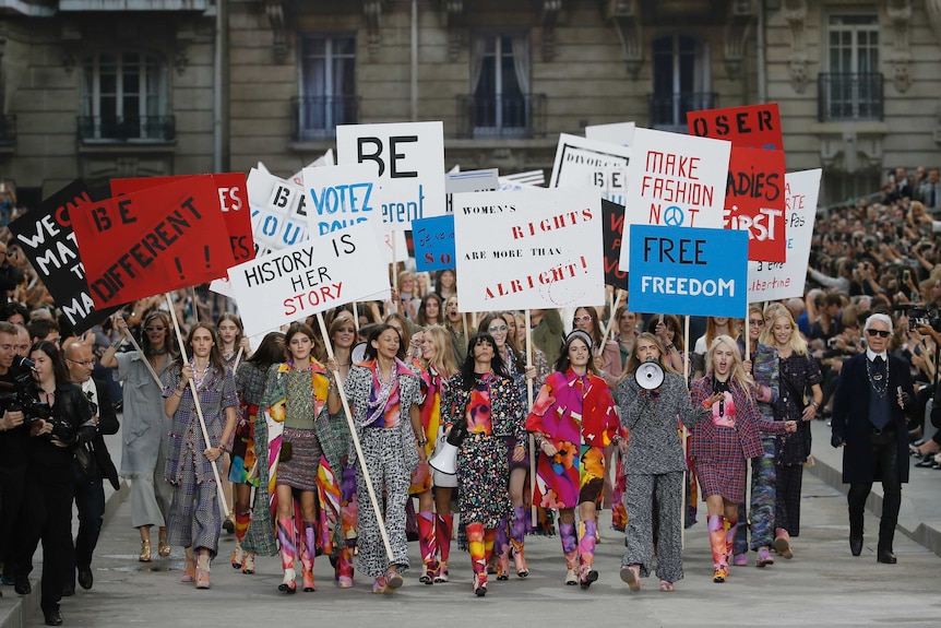 A large group of models walk down a runway holding placards with various slogans and words on them.