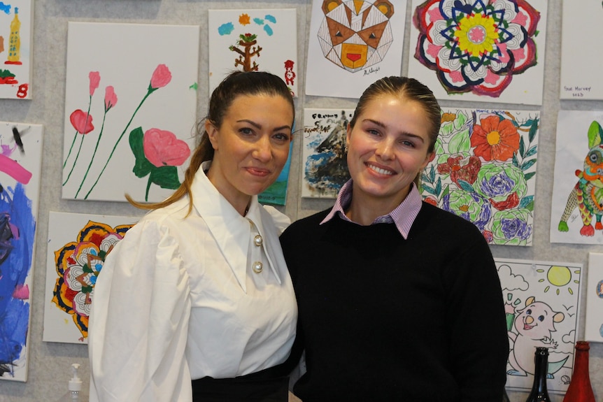 Two women smile at the camera in front of a wall of children's drawings