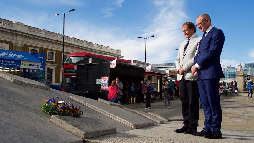 Two men dressed in suits stand solemnly looking at a floral wreath laid on the edge of a bridge.