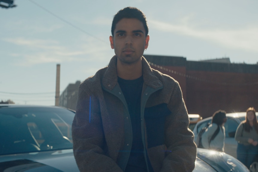 actor rish shah during a scene of ms marvel. he is leaning on the bonnet of a car wearing a black t shirt and brown jacket