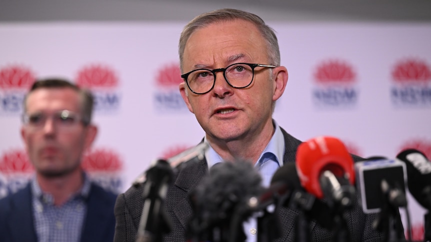 Anthony Albanese looks serious, speaking into a row of microphones, with Dominic Perrottet out of focus behind him