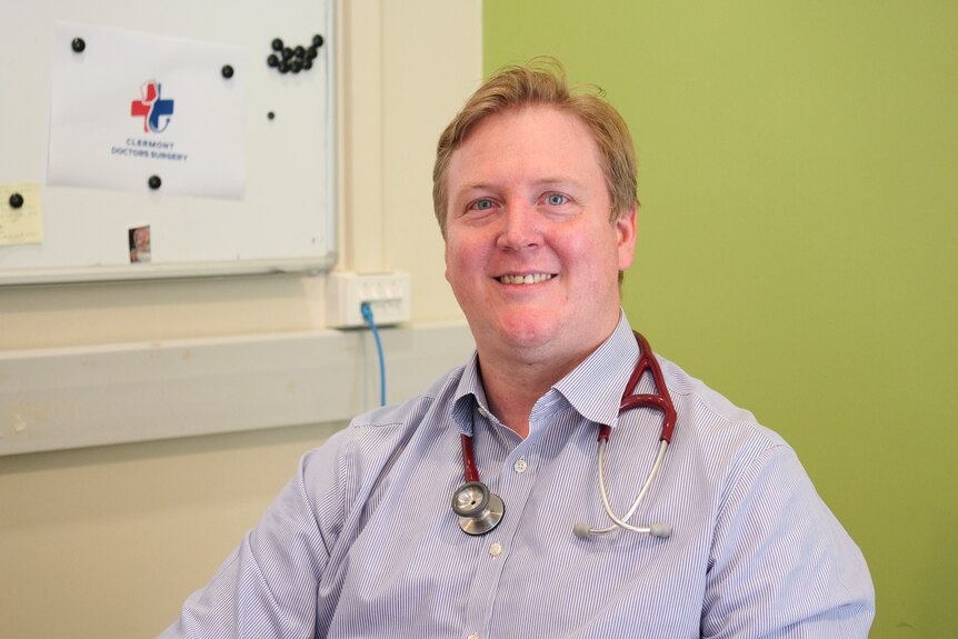 A doctor sits at his desk with a red stethoscope around his neck in a blue button up shirt with a green wall and whiteboard