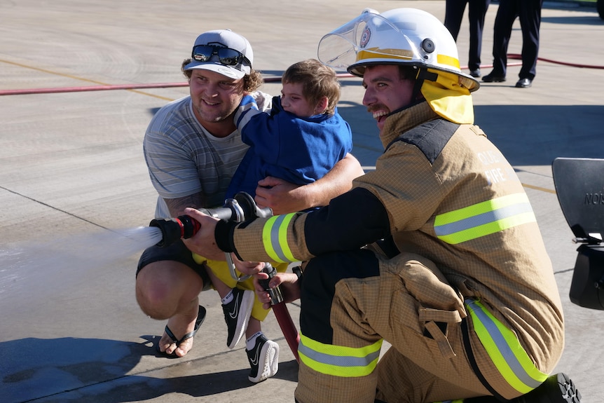 a firefighter kneels down with a man and boy to use a fire hose