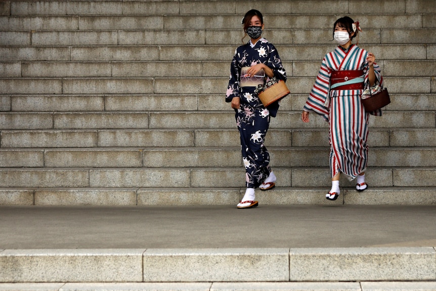 Two women wearing kimonos and masks walk down steps. 