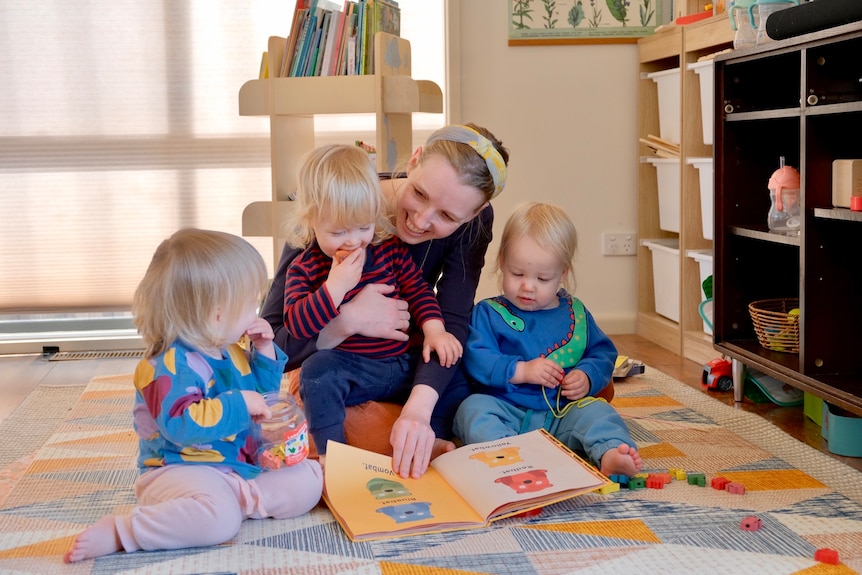 A woman sits on the floor with her three children reading a book.