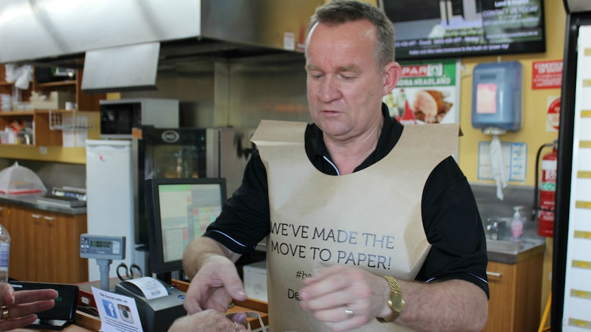 Man stands serving a customer at a supermarket, handing bag change, wearing a paper bag vest.