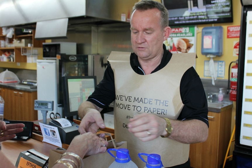 Man stands serving a customer at a supermarket, handing bag change, wearing a paper bag vest.
