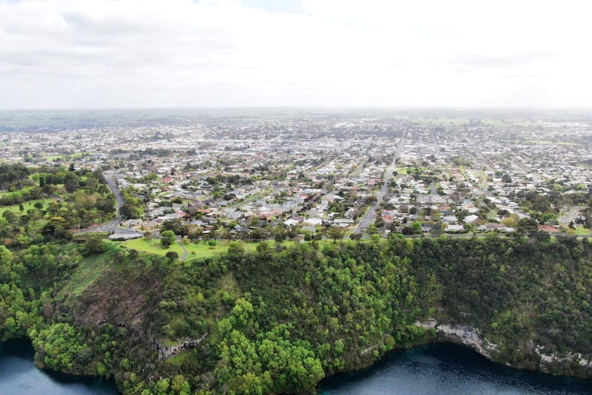 An aerial photo of a large grassy, tree-lined cliff sloping into a large blue lake, rows of houses and roads behind it.