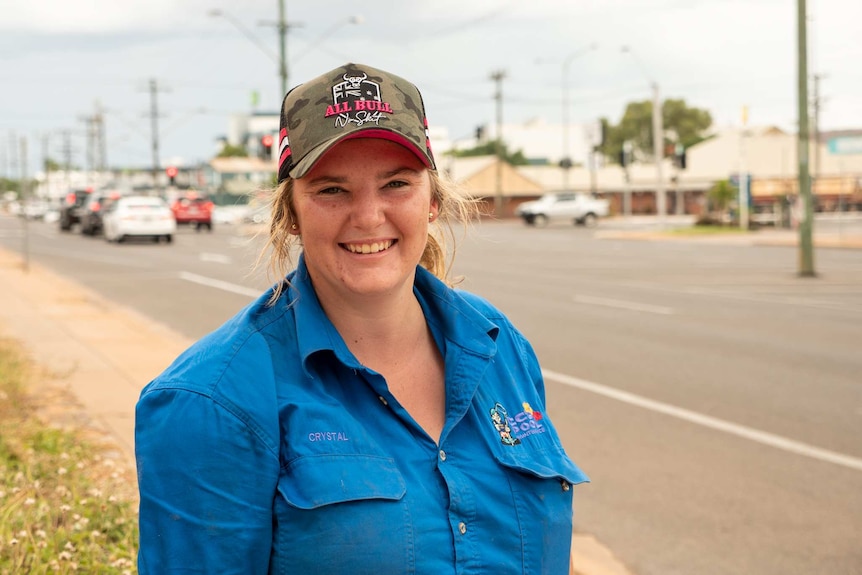 A woman wearing a cap and blue work shirt stands on a footpath, she is smiling.